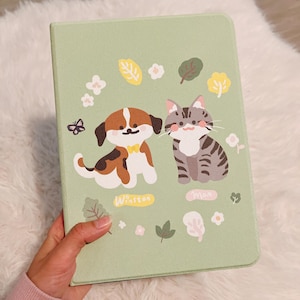 Könnte beinhalten: Ein grüner Notizbuchumschlag mit einer niedlichen Illustration eines Hundes und einer Katze. Der Hund ist braun und weiß mit schwarzen Flecken und die Katze ist grau und weiß mit schwarzen Streifen. Die Illustration ist von Blumen und Blättern umgeben. Der Text "Winston" und "Max" steht unter dem Hund und der Katze.