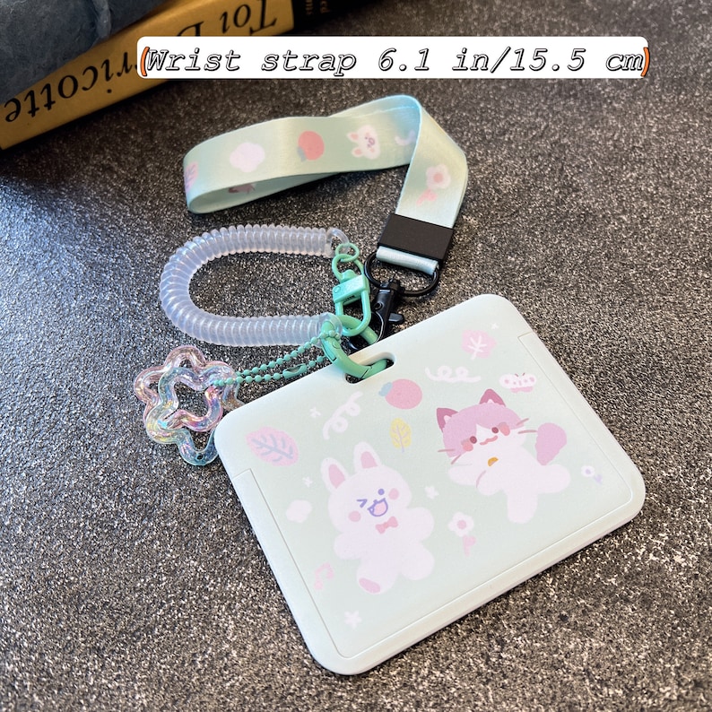 Op de afbeelding: Een lichtgroene rechthoekige kaarthouder met een print van een wit konijn en een roze kat. Het bevat een polsband van 15,5 cm, een transparant spiraalsnoer en een doorschijnende, iriserende stervormige bedel bevestigd aan een balletjesketting.