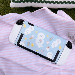 May include: A handheld gaming console featuring a light blue protective skin decorated with white cartoon seals, ice cream cones, and boba tea drinks. The device has white side grips and black buttons, showcasing a playful pattern of treats.