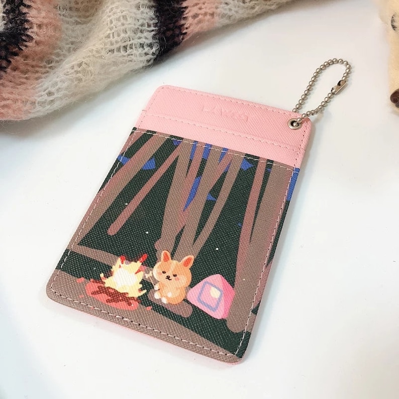 May include: A rectangular card holder featuring a pink top section and a graphic print of a corgi sitting by a campfire in a forest at night. The accessory includes a silver-toned metal ball chain attached to the top right corner for hanging.