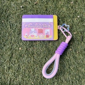 May include: A purple and yellow card holder with a cartoon illustration of a cat in a baker's hat. The card holder has a pink and white lanyard with a star-shaped charm. The card holder has the text "Starry Forest Bakery" printed on it.