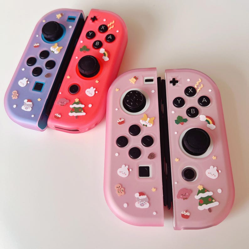 May include: Three colorful Nintendo Switch Joy-Con controllers. One is lavender, one is red, and one is pink. Each controller has black buttons and a black joystick. The controllers are decorated with various holiday-themed stickers.