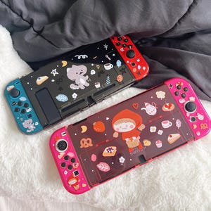 Może przedstawiać: Dwa futerały na gry Nintendo Switch z kolorowymi wzorami. Górny futerał jest czarny z białym i szarym wzorem kota, a dolny futerał jest przezroczysty z różowym i czerwonym wzorem przedstawiającym dziewczynkę w czerwonej sukience z kapturem i różnymi innymi uroczymi ilustracjami.