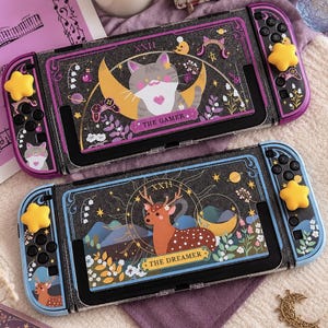 Może przedstawiać: Dwie przenośne konsole do gier z etui ochronnymi w stylu tarota. Górna konsola przedstawia kota na półksiężycu z napisem THE GAMER. Dolna konsola pokazuje jelenia w lesie z napisem THE DREAMER. Obie mają żółte nakładki na joysticki w kształcie gwiazdek.