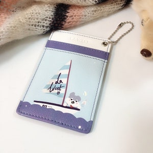 May include: A rectangular card holder featuring a light blue background with a graphic of a sailboat and a small character. The design includes purple scalloped trim at the bottom and a purple band at the top. The text "La West" is printed on the sail, and a silver metal ball chain is attached to the top right corner.