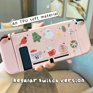 K&ouml;nnte beinhalten: Rosa Nintendo Switch H&uuml;lle mit Winter-Weihnachts-Motiv. Die H&uuml;lle zeigt einen Schneemann, einen Lebkuchenmann und andere festliche Designs. Der Text "All TPU soft material" und "Regular Switch version" ist auf der H&uuml;lle aufgedruckt.