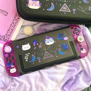 May include: A black and purple Nintendo Switch console with a matching carrying case. The console and case are decorated with cat and celestial-themed stickers. The case has the text "Starry Forest Divination".
