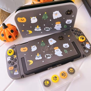 May include: A gray Nintendo Switch console and case decorated with Halloween-themed stickers. The stickers include pumpkins, ghosts, Christmas trees, and the word "Boo". Additional paw-print and pumpkin-themed thumb grips are also visible.