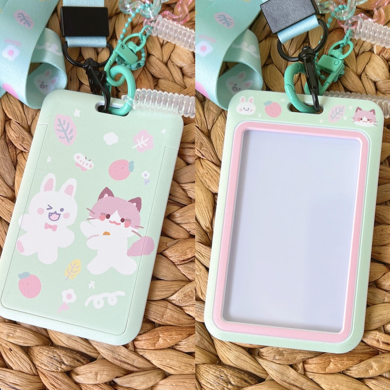 Op de afbeelding: Een lichtgroene verticale badgehouder met een cartoonillustratie van een konijn en een kat met roze en witte accenten. De houder is voorzien van een bijpassend lichtgroen koord met een doorzichtige plastic spiraal en een zwarte clip.