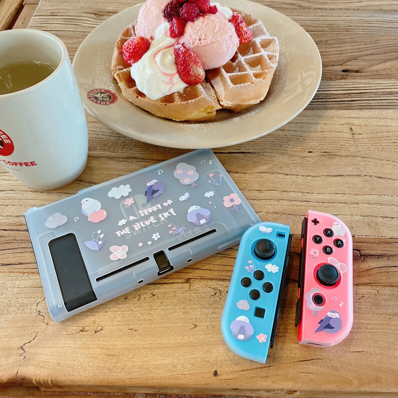 K&ouml;nnte beinhalten: Ein hellgraues Nintendo Switch-Geh&auml;use und Controller mit einem Thema des blauen Himmels. Das Geh&auml;use und die Controller zeigen Illustrationen von V&ouml;geln, B&auml;umen und Blumen. Der Text "A Story of the Blue Sky" ist auf dem Geh&auml;use aufgedruckt. Eine Waffel mit Eis und Erdbeeren liegt auf einem Teller.