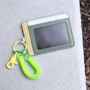 May include: A green, yellow, and white leather ID card holder with a clear plastic window. A yellow and green retractable key chain is attached to the holder.
