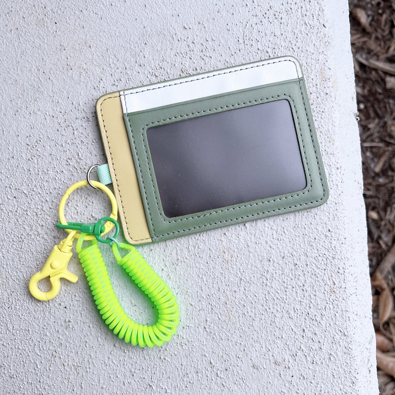 May include: A green, yellow, and white leather ID card holder with a clear plastic window. A yellow and green retractable key chain is attached to the holder.