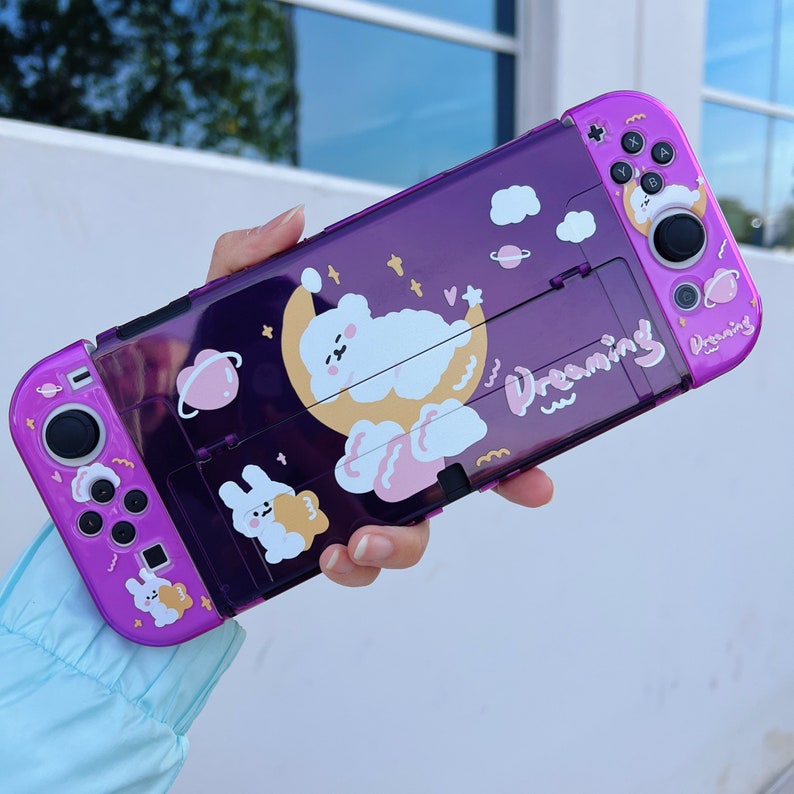 May include: Purple clear plastic case for a Nintendo Switch with a cute cartoon bear design and the word "Dreaming" printed on it.