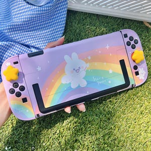 May include: A lavender Nintendo Switch case featuring a cartoon bunny, rainbow, and stars. The case has yellow star-shaped button covers and the words "Starry Forest". The design is on a purple background.