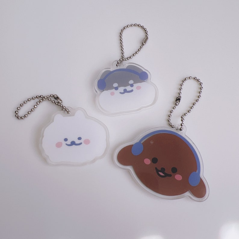 Op de afbeelding: Drie sleutelhangers met cartoon dierenfiguren die een koptelefoon dragen. De figuren zijn een witte hond, een bruine hond en een grijze hond. Elke sleutelhanger is gemaakt van helder acryl en heeft een zilveren ketting.