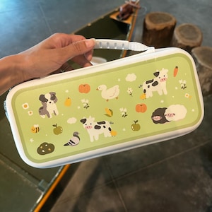 以下が含まれることがあります： 白のハンドルとトリムが付いた、長方形の明るい緑色のケース。犬、牛、アヒル、羊などの家畜、カボチャ、リンゴ、花が繰り返し描かれたパターンが特徴です。ケースは白いジッパーで閉じられています。