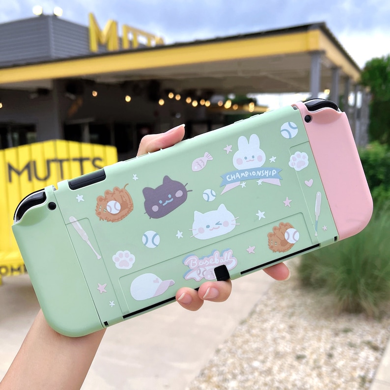 May include: A light green Nintendo Switch case with a pink Joy-Con controller. The case is decorated with cartoon animal stickers, including a cat, a rabbit, and a baseball. The text "Baseball Championship" is printed on the case.