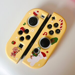 May include: Yellow Nintendo Switch Joy-Con controllers with festive holiday-themed designs. The controllers feature black buttons, a directional pad, and various decorative stickers including a cupcake, musical notes, and a candy cane.