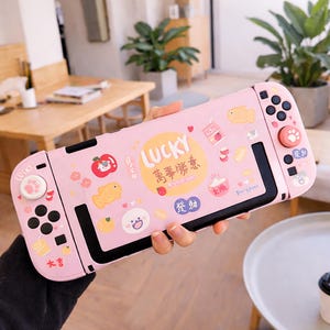 Könnte beinhalten: Rosa Nintendo Switch-Hülle mit Cartoon-Illustrationen, darunter ein Fisch, ein Apfel und das Wort "LUCKY". Die Hülle hat schwarze Akzente und rosa Knöpfe. Das Design enthält japanische Schriftzeichen und einen Pfotenabdruck.