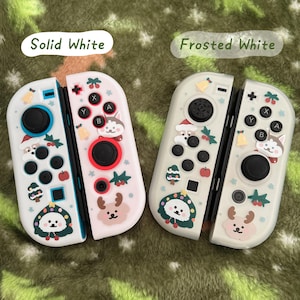 May include: Two white Nintendo Switch Joy-Con controllers. One is solid white, the other is frosted white. Both are decorated with festive holiday-themed graphics, including a reindeer, a husky, and a Christmas tree. The controllers have black buttons and red and black accents.