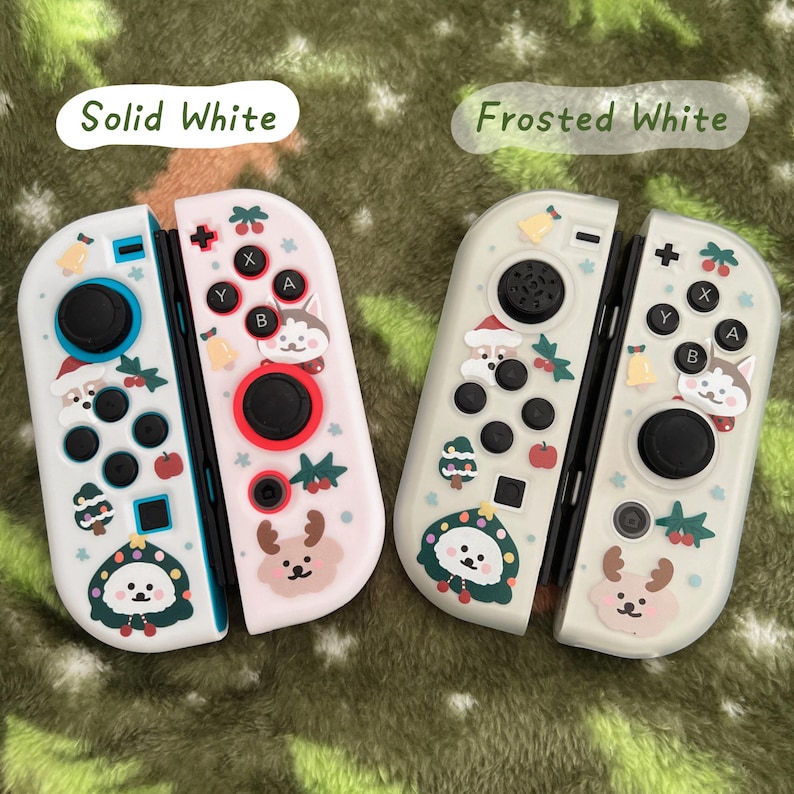 May include: Two white Nintendo Switch Joy-Con controllers. One is solid white, the other is frosted white. Both are decorated with festive holiday-themed graphics, including a reindeer, a husky, and a Christmas tree. The controllers have black buttons and red and black accents.