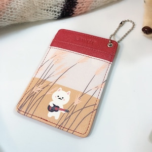 May include: A rectangular card holder featuring a red top section and a light-colored lower section with a graphic of a white cat playing a small guitar amidst tall grass. A silver metal ball chain is attached to the top right corner.