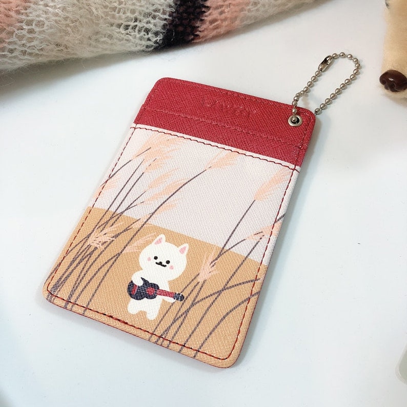 May include: A rectangular card holder featuring a red top section and a light-colored lower section with a graphic of a white cat playing a small guitar amidst tall grass. A silver metal ball chain is attached to the top right corner.