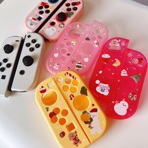 May include: A collection of colorful Nintendo Switch Joy-Con controllers and protective cases. The controllers are white, pink, and yellow, with various Christmas-themed designs, including snowmen, Santa, and Christmas trees. The cases are transparent and decorated with festive stickers.