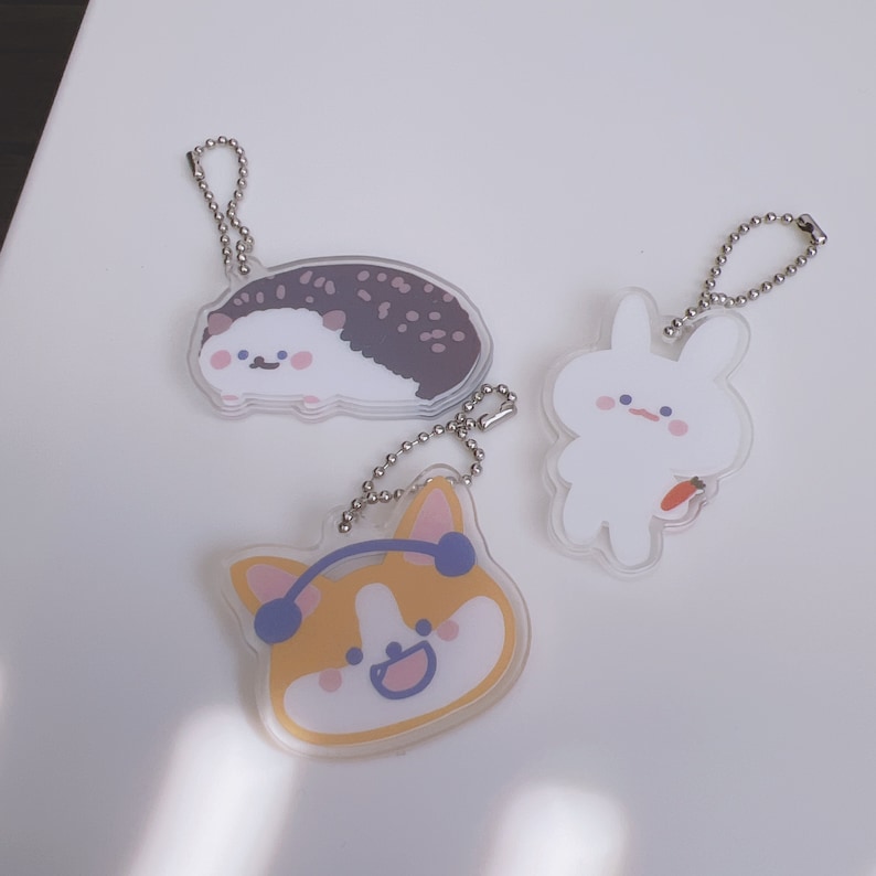 Op de afbeelding: Drie sleutelhangers met cartoon-dierontwerpen. De eerste sleutelhanger is een bruin-witte egel, de tweede is een wit konijn dat een wortel vasthoudt, en de derde is een gele en oranje corgi met een koptelefoon.