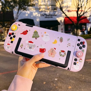 Puede incluir: Una funda rosa para Nintendo Switch adornada con pegatinas con temática navideña. La funda presenta una variedad de diseños navideños, que incluyen un muñeco de nieve, un hombre de jengibre y un árbol de Navidad. Los botones son amarillos y rosas.