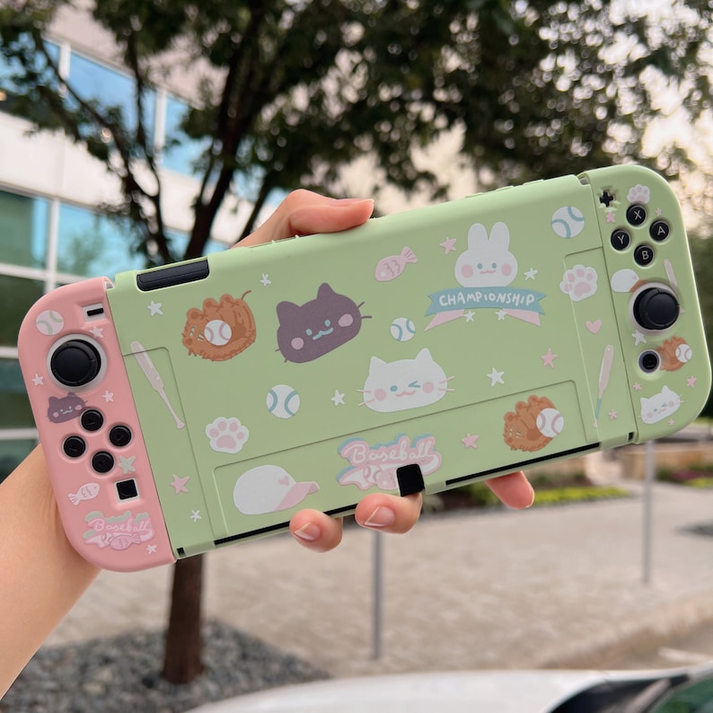 May include: A green and pink Nintendo Switch case with a baseball theme. The case features a variety of cute cartoon characters, including cats, a bunny, and a baseball. The text "Baseball Championship" is printed on the case.