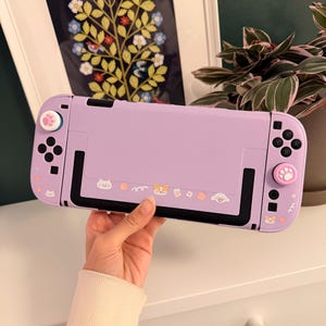 May include: A lavender Nintendo Switch console with a protective case. The case features black accents, paw print thumb grips, and decorative stickers of cats and other cute characters. The console is held by a person.