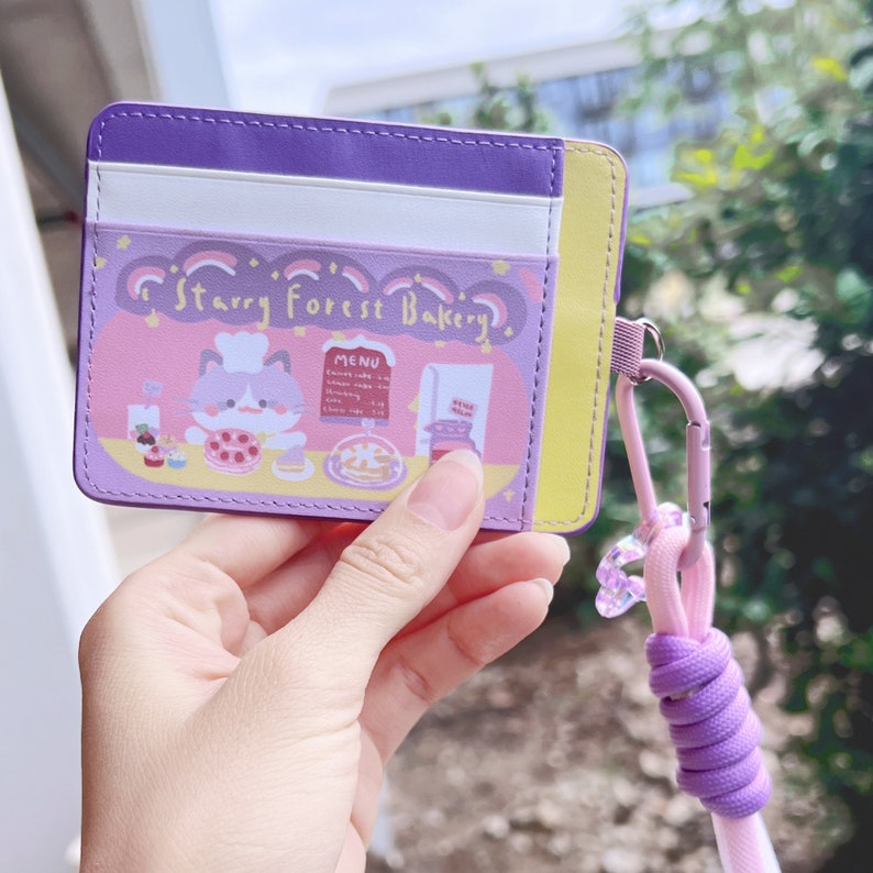 May include: Purple card holder with a cartoon cat wearing a chef's hat and a sign that says "Starry Forest Bakery". The card holder has a purple lanyard with a clear charm.
