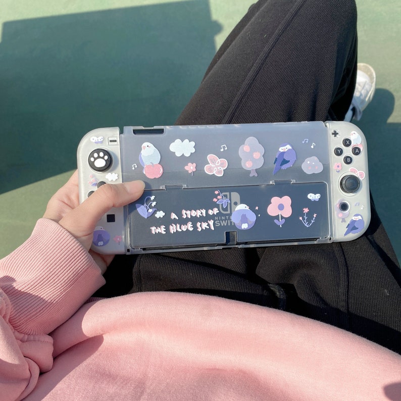 K&ouml;nnte beinhalten: Eine graue Nintendo Switch-Konsole mit einer transparenten Schutzh&uuml;lle. Die H&uuml;lle ist mit Aufklebern von V&ouml;geln, Blumen und B&auml;umen verziert. Der Text "A Story of the Blue Sky" ist auf der H&uuml;lle aufgedruckt.