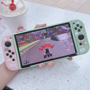 May include: A green and pink Nintendo Switch console with a racing game displayed on the screen. The game features a cartoon character driving a kart on a track with red balloons. The console has a pink Joy-Con controller on the left and a green Joy-Con controller on the right.