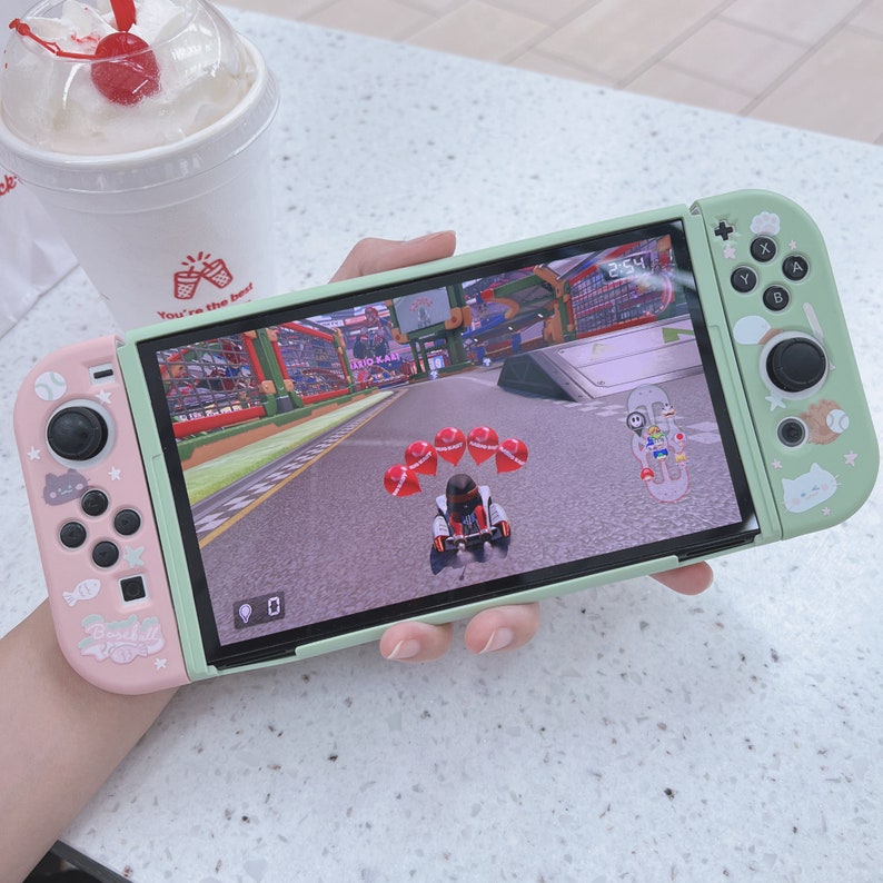 May include: A green and pink Nintendo Switch console with a racing game displayed on the screen. The game features a cartoon character driving a kart on a track with red balloons. The console has a pink Joy-Con controller on the left and a green Joy-Con controller on the right.
