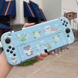 Op de afbeelding: Een lichtblauwe Nintendo Switch-hoes met een bloemenontwerp met witte bloemen, groene bladeren en kleine witte bellen. De hoes heeft ook cartoon-dierillustraties van een hond, een beer en een konijn. De tekst "Spring Time" staat op de hoes gedrukt.