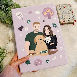 Puede incluir: Un cuaderno ilustrado morado con una pareja, un perro y un gato. El cuaderno tiene un diseño floral con un árbol, flores y bayas. El texto en el cuaderno dice "Jacob, Charlie, Olivia".