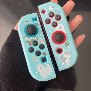 May include: A pair of light blue Nintendo Switch Joy-Con controllers. Each controller features black buttons and control sticks, with decorative stickers of a snowman, husky, and other winter-themed designs.
