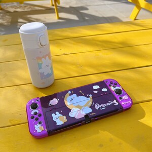 May include: A purple Nintendo Switch console with a white and pink cartoon design. The console has a white and pink cartoon design with a cloud, stars, and the word "Dreaming". The console is sitting on a yellow table next to a white water bottle with a pink and white cartoon design.