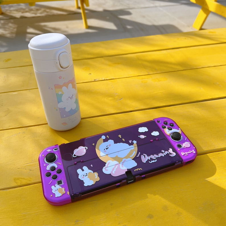 May include: A purple Nintendo Switch console with a white and pink cartoon design. The console has a white and pink cartoon design with a cloud, stars, and the word "Dreaming". The console is sitting on a yellow table next to a white water bottle with a pink and white cartoon design.