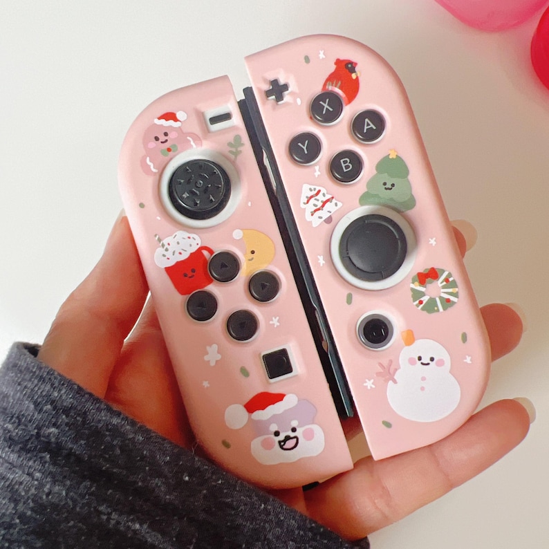 May include: Pink Nintendo Switch Joy-Con controllers decorated with Christmas-themed stickers. The stickers include a gingerbread man, a snowman, a cardinal, a Christmas tree, and a mug of hot cocoa.