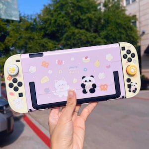 May include: A light purple Nintendo Switch case with a cute panda and cat design. The case has a black frame and yellow accents on the controllers. The case is decorated with food and musical note graphics.