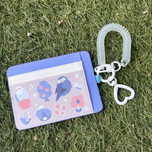 May include: A purple card holder with a white and pink floral design featuring birds and musical notes. The card holder has a white strap with a clear plastic coil.