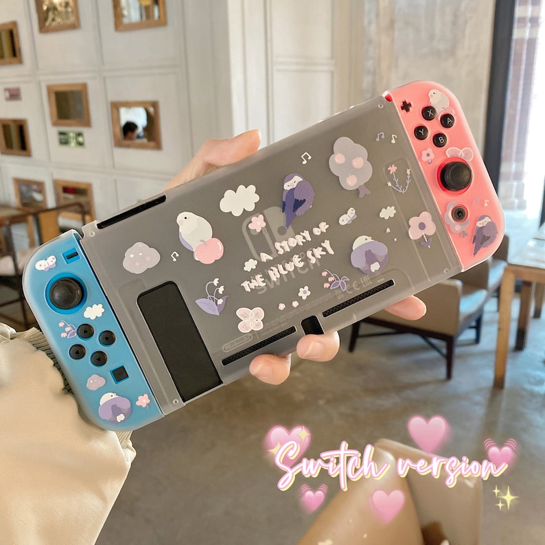 K&ouml;nnte beinhalten: Eine Nintendo Switch-Konsole mit einer grauen, transparenten H&uuml;lle mit Illustrationen von V&ouml;geln, Wolken und Blumen. Der Text "A Story of the Blue Sky" ist auf der H&uuml;lle aufgedruckt. Die Controller sind blau und rosa.
