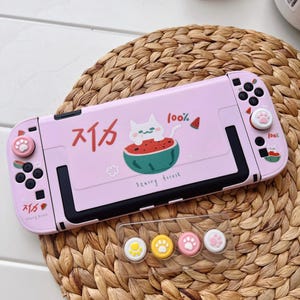 May include: A pink Nintendo Switch case with a cat and watermelon design. The case has Japanese text and the words "Starry Forest". Includes matching thumb grips with paw prints. The case is on a woven surface.