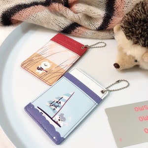 May include: Two rectangular card holders with metal ball chains. One is red and orange with a white cat playing a guitar in tall grass. The other is light blue and purple with a sailboat graphic and the text "la west" featuring a small character.