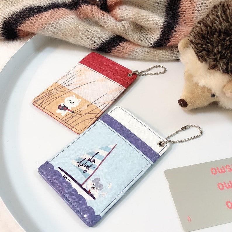 May include: Two rectangular card holders with metal ball chains. One is red and orange with a white cat playing a guitar in tall grass. The other is light blue and purple with a sailboat graphic and the text "la west" featuring a small character.