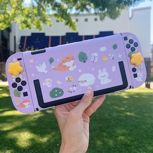 May include: A lavender-colored gaming console case with a whimsical design. The case features cartoon illustrations of a cat, bunny, sheep, and other cute elements. Yellow star-shaped buttons and black control buttons are visible.