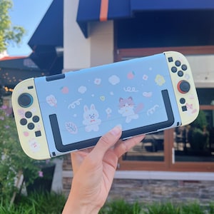 May include: A light blue Nintendo Switch case with a whimsical design featuring a white bunny, a fox, and other cute illustrations. The case has yellow accents on the sides and buttons, and a black frame around the screen.